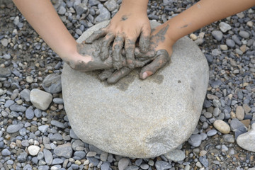 Manos de niño, una sobre otra, con barro, sobre una piedra.