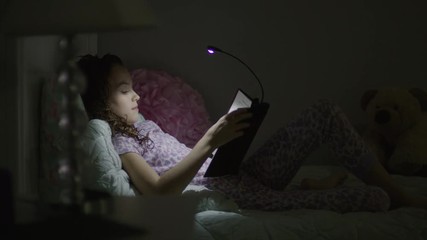 Medium shot from window of girl reading book in bed at night / Provo, Utah, United States