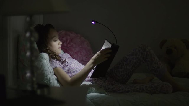 Medium shot from window of girl reading book in bed at night / Provo, Utah, United States