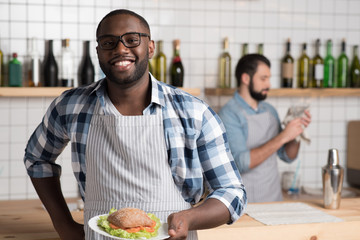 Beautiful meal. Emotional handsome experienced waiter standing at the bar counter and holding a plate with a big tasty sandwich while his colleague cleaning the classes behind his back