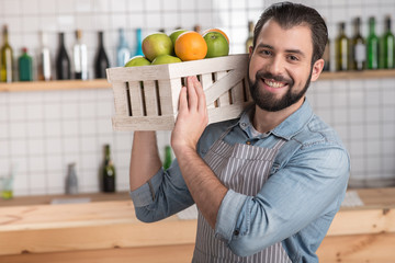 Fresh fruit. Handsome responsible enthusiastic worker looking not tired while holding a heavy wooden box with tasty fresh fruit and smiling cheerfully