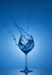 Water splash in out of glass. Water splashing out of a tall wine glass. Blue gradient background. Reflection.