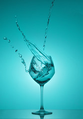 Water splash in out of glass. Water splashing out of a tall wine glass. Turquoise gradient background. Reflection.