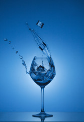 Ice cubes fall into a glass and water is poured out. Water splashing out of a tall wine glass. Gradient background and reflection.