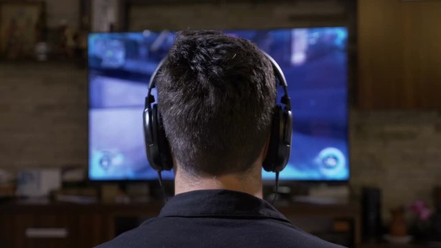 Captivated young man player with big earphones relaxing at home gaming in front of digital monitor TV