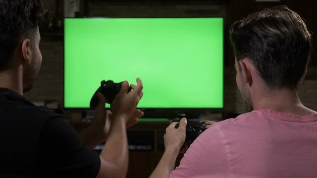 Young friends having fun with wireless joysticks competing with each other playing video games on chroma key green screen
