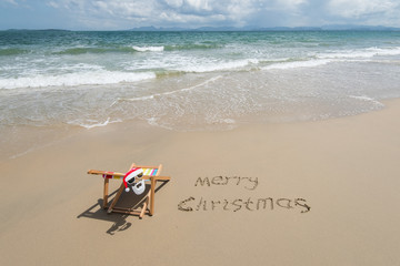 Merry Christmas written in sand write on tropical beach.Deck chair with Santa Claus sunglasses