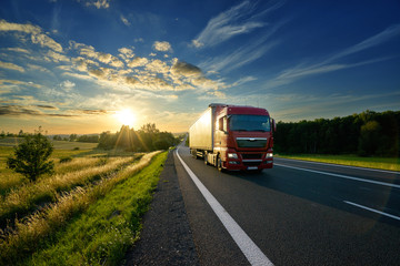 Fototapeta na wymiar Red truck driving on the asphalt road in rural landscape at sunset