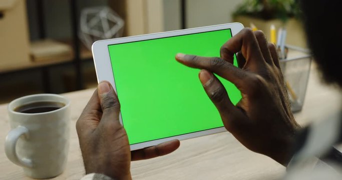 Over shoulder view on the white tablet computer holded by african american man horizontally. Fingers scrolling and taping on it. Green screen. Chroma key. Wooden office desk with cup of coffee