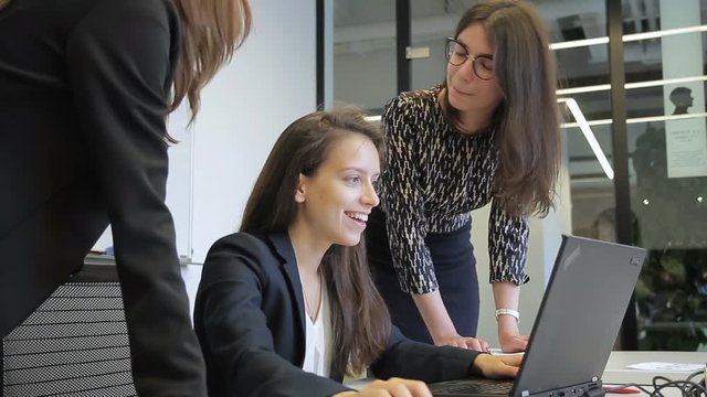 In office three young women have fun in front of laptop on desktop. Female employees in business outfit are happy and have a nice time near screen during break.