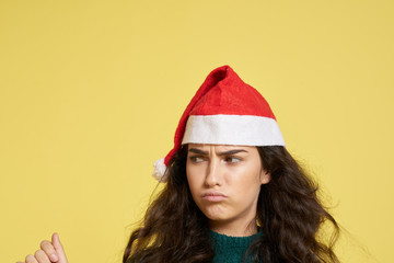 light yellow background, free space for copy, female brunette in a Christmas hat puffs up her lips and frowns, new year