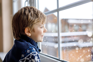 Happy adorable kid boy sitting near window and looking outside on snow on Christmas day or morning.