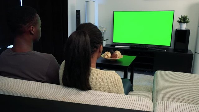 Young black couple watch television in living room and then smile to camera 