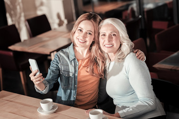 Happy together. Kind pleasant cheerful woman looking glad while sitting in a cafe and hugging her beautiful elderly granny