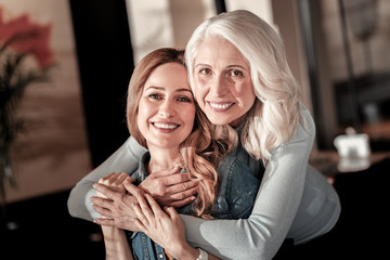 Huggins relatives. Cheerful emotional kind senior woman looking glad while hugging her pretty positive young granddaughter