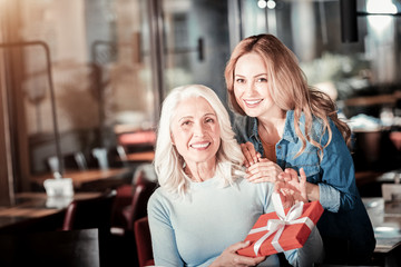 Little holiday. Cheerful glad senior woman sitting at the table and showing a red box with a nice present from her kind loving granddaughter