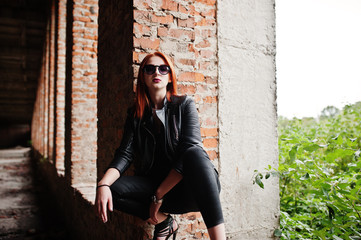 Red haired stylish girl in sunglasses wear in black, against abadoned place with brick walls.