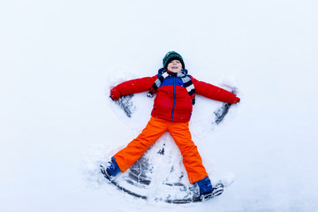 Cute little kid boy in colorful winter clothes making snow angel, laying down on snow