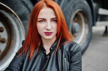 Plakat Red haired stylish girl wear in black, sitting against large truck wheels.