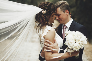 Groom hugs bride tender while wind blows her veil somewhere in Tuscany, Italy