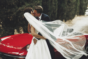 Wind blows bride's veil while groom hugs her standing before a red retro cabriolet
