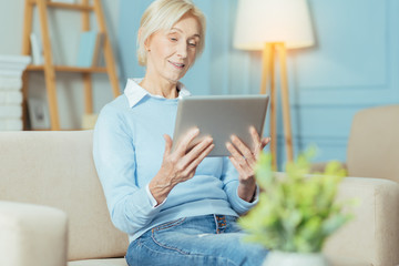 Curious lady. Smart enthusiastic senior woman feeling progressive while staying at home and looking at the screen of a modern tablet while watching an interesting film