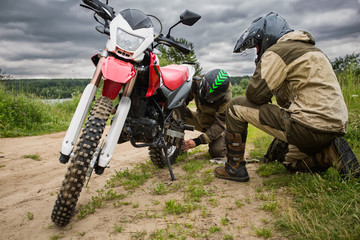 Two men in motorcycle gear checking rear wheel of dirtbike.