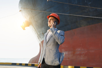 Handsome young unshaven successful business man in gray suit and protective construction orange helmet holding tablet, talking on mobile phone, standing in sea port against cargo rusty ship background
