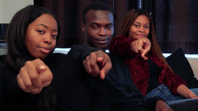 Three african friends point to camera with finger and smile in living room