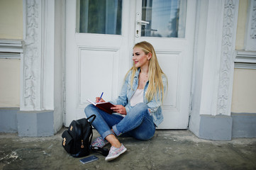 Blonde girl wear on jeans with backpack posed against old door with diary and write something.