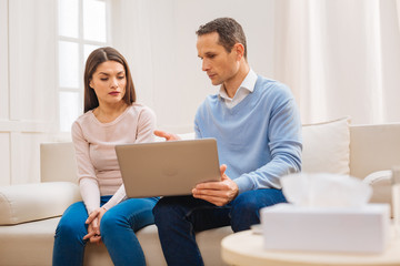 Our future. Attractive young focused couple sitting on the sofa and looking at the screen while man holding laptop 