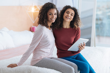 No time for worries. Joyful young ladies sitting on the edge of a bed with cheerful smile on their faces and looking into the camera while working on a touchpad together.