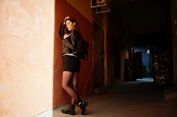 Stylish brunette girl wear on leather jacket and shorts with backpack against orange wall at shadows.