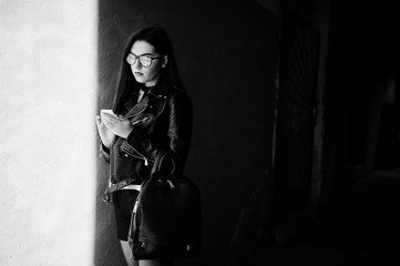 Stylish brunette girl wear on leather jacket and shorts with backpack and glasses against orange wall at shadows looking on mobile cell phone.