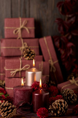 Three candles of crimson and pink color on the background of gift boxes with cones, leaves and daisies.