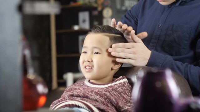 Barber in black gloves makes a hairstyle for an Asian child, puts a gel on his hair 60 fps