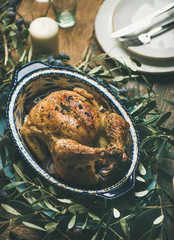 Whole roasted chicken for Christmas eve celebration decorated with olive tree branch, plates and candles over rustic wooden table background. Holiday table decoration set concept