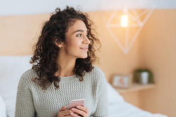 What if. Thoughtful young lady smiling while looking into vacancy with her smartphone in hands and thinking of something pleasant.