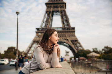 Beautiful girl posing to the photographer against the background of the Eiffel Tower. Autumn photosession. Sunny weather. Beautiful smile and makeup