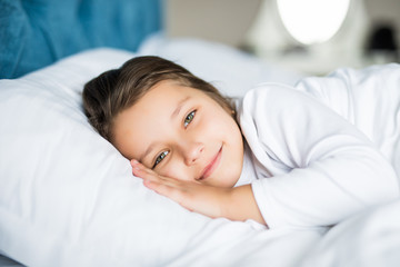 Fototapeta na wymiar Close-up portrait of laughing little girl lying on bed with hand under pillow, looking at camera and smile. Good morning.