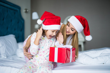 Happy family wearing Santa Claus hats sitting on bed. Mother give and surprised child Santa gift and having fun in Christmas time. Winter holiday Xmas and New Year concept