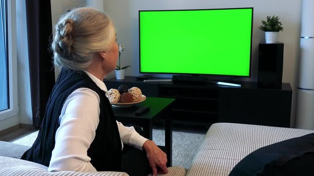 Old caucasian woman watches television in living room and then agrees to camera 