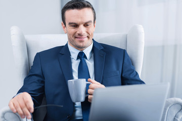 Routine work. Attractive nice young man  sitting in the armchair and drinking tea while looking at the screen