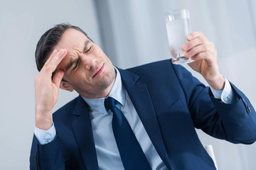 Headache driving crazy. Sad brunette young man closing his eyes while carrying glass of water and touching his forehead 