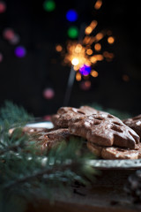 gingerbread cookies for the new year on a silver tray, New Year's lights, fireworks, fur-tree, cones, cinnamon on a wooden cutting board on a dark background