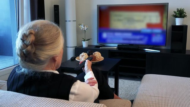 Old caucasian woman watches television in living room and switches program 