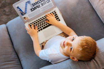 Obraz na płótnie Canvas Smart child. Top view of a cute pleasant young boy sitting on the sofa and looking at you while holding his laptop