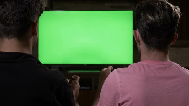 Couple of chilling focused guys on sofa playing video game with joypads on chroma key green TV screen