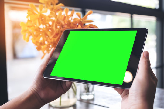 Mockup image of hands holding black tablet pc with blank green screen and flower vase on wooden table in cafe
