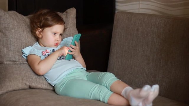 Funny child looks at the phone screen and plays downloaded application on a smart phone close-up. A little cute girl lies in sofa in a living room, looking cartoon and playing the game